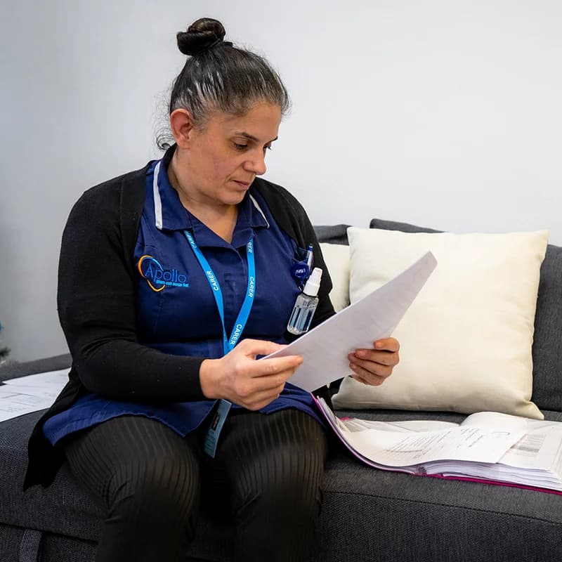 Apollo Care carer in branded blue uniform and lanyard reviewing a client's care-plan paperwork on a home sofa during a domiciliary visit