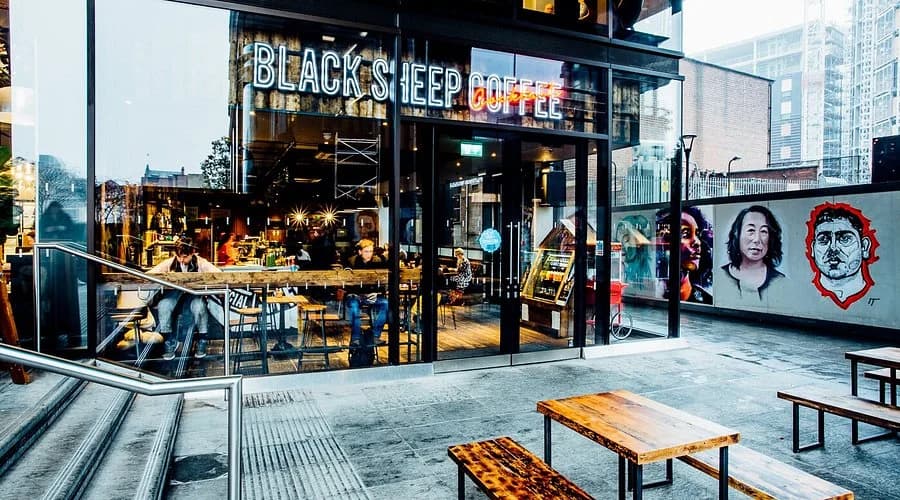 Black Sheep Coffee storefront with full-glass facade, neon signage, outdoor seating and street-art portraits on the adjacent wall