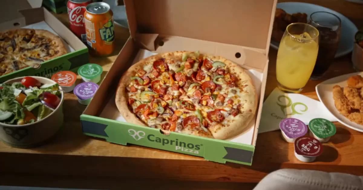 Caprinos Pizza branded takeaway box with a freshly cooked pizza inside, served alongside a side salad, soft drinks and dipping sauces - a typical Caprinos meal-deal at home