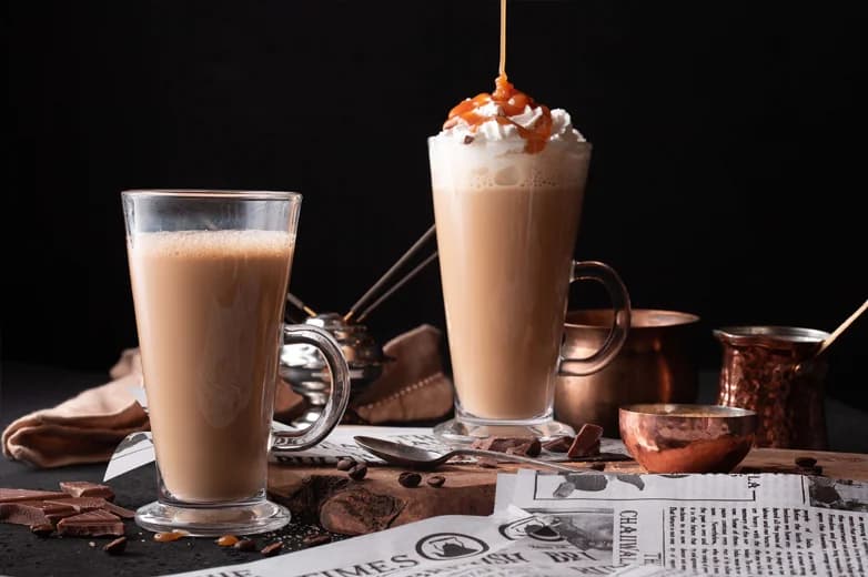 Chaiiwala signature karak chai latte in tall glass mugs - one with whipped cream and caramel drizzle, served alongside traditional copper chai pots