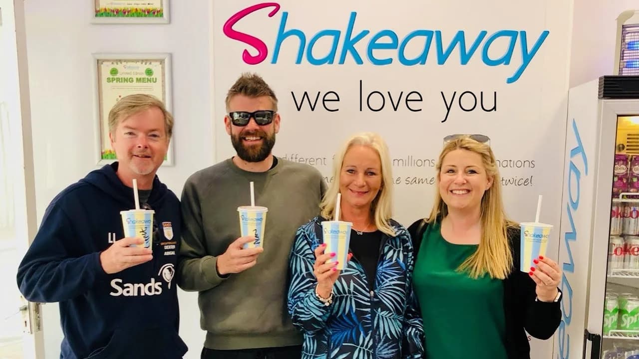 Four happy Shakeaway customers holding their milkshakes in front of a 'Shakeaway we love you' branded wall
