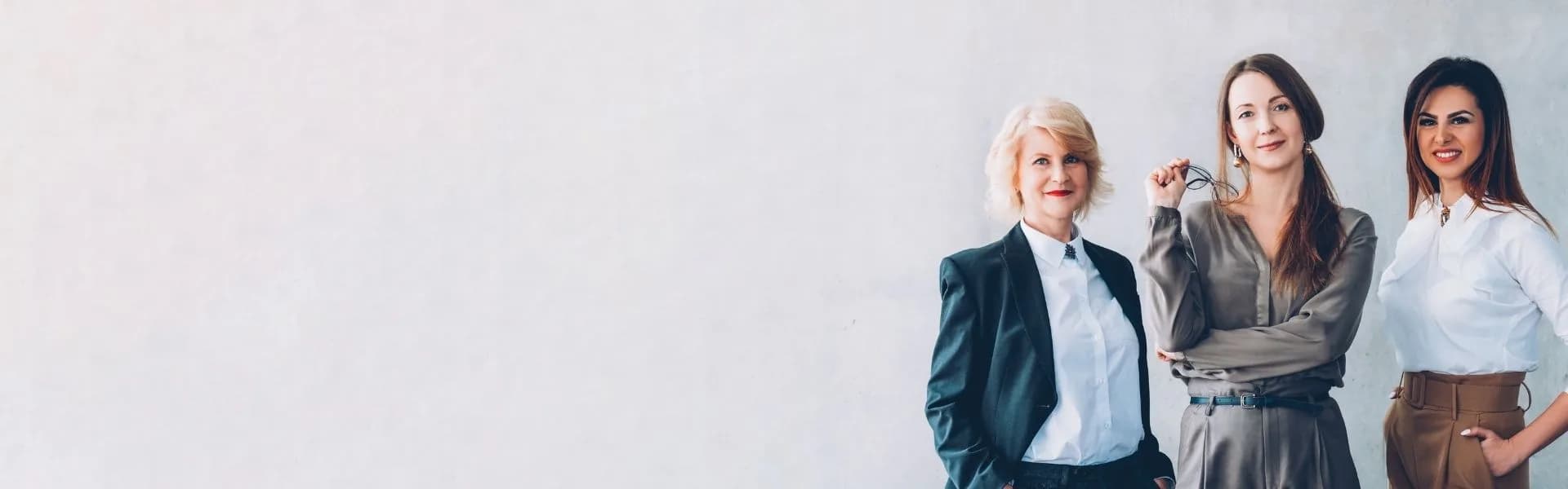 Three confident businesswomen in smart professional attire standing together against a light grey wall, representing full-time franchise opportunities in the UK.