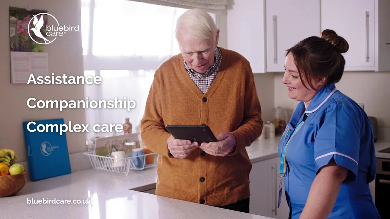 Bluebird Care nurse in blue uniform showing a tablet to a smiling elderly client in his home kitchen, with text overlays for the brand's Assistance, Companionship and Complex care services