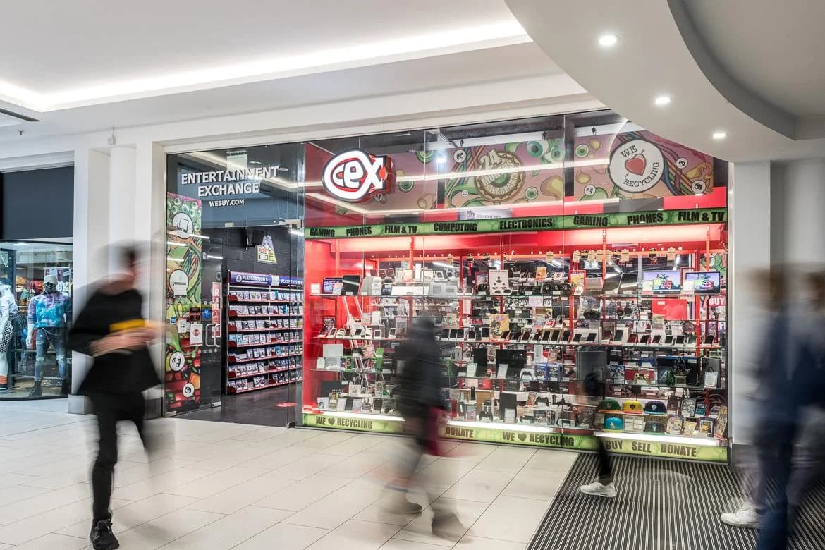CeX shopping-mall shopfront with red 'Entertainment Exchange' signage, 'WeBuy.com' branding and product departments for Gaming, Phones, Film and TV, Computing and Electronics on display inside