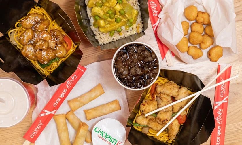 Aerial flat-lay of a full Chopstix Noodle Bar menu spread - salt and pepper chicken noodle box, vegetable curry rice with peas, popcorn chicken, salt and pepper chicken wok box, BBQ pork ribs, spring rolls and branded 'Click your sticks' chopsticks on a wooden table