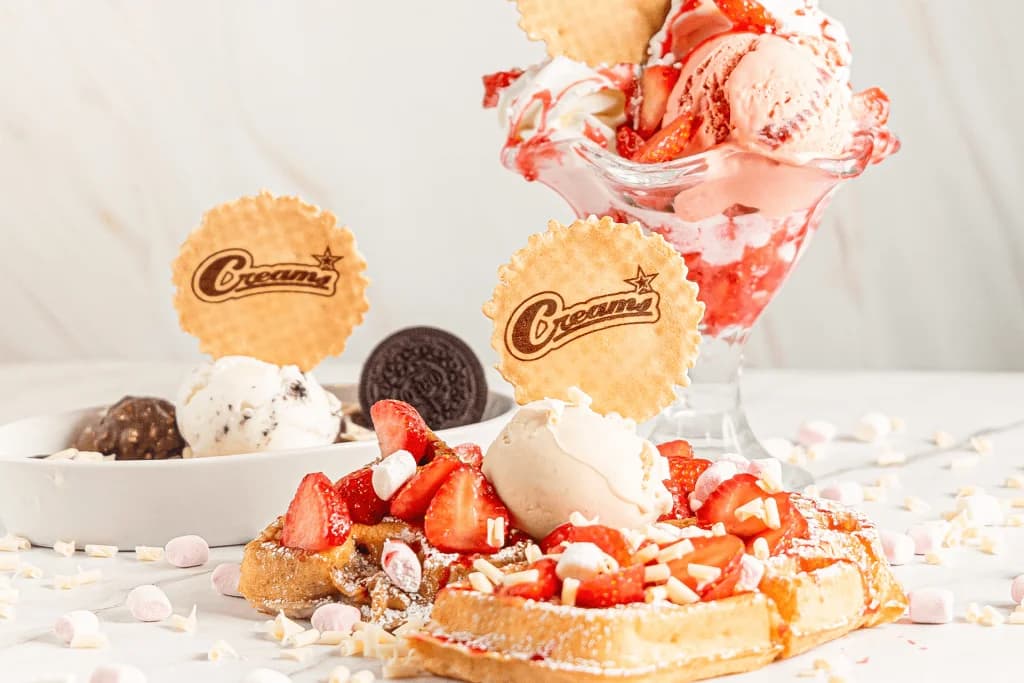 Creams Cafe close-up plate: a Belgian waffle topped with fresh strawberries, vanilla ice cream and cream, paired with a strawberry-gelato sundae glass, Oreo crumb plate and branded Creams wafers