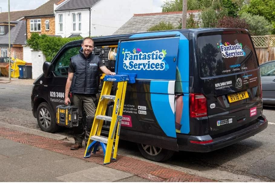 Fantastic Services franchisee in a branded vest standing beside his wrapped VW Transporter Fantastic Services van, with a yellow extension ladder and a black tool case ready for the day's home services jobs