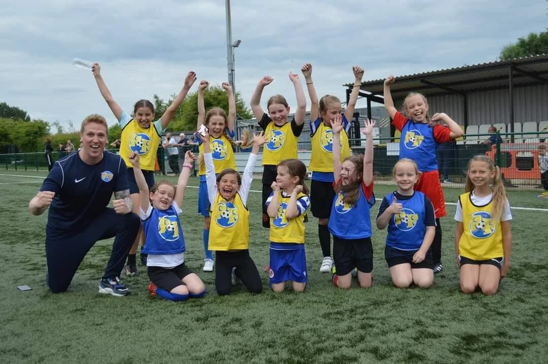 A Football Fun Factory coach in branded training jacket kneeling alongside a group of girls aged 6-10 in blue and yellow Football Fun Factory bibs, all cheering with arms raised on a 3G astroturf pitch after a session