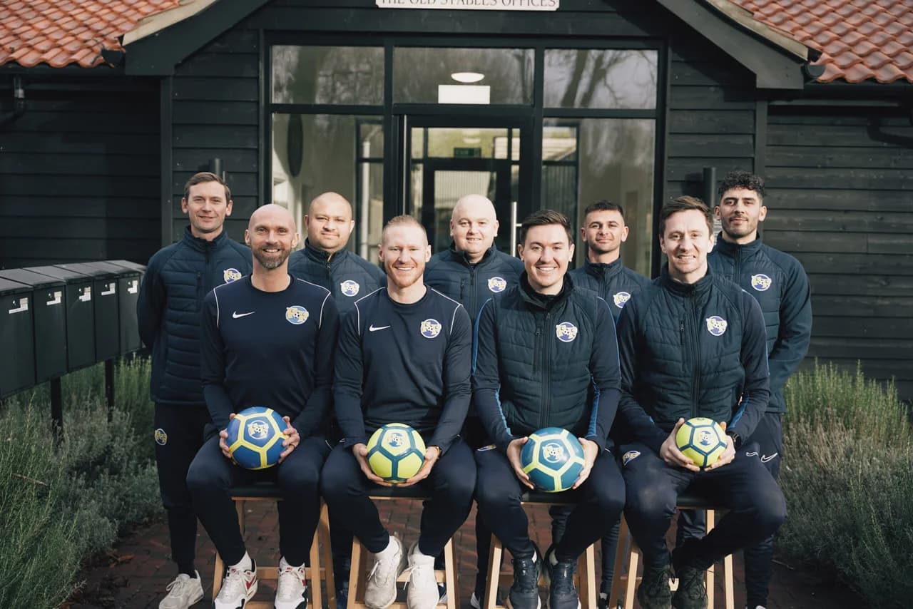 The Football Fun Factory head office team - eight coaches in matching navy training jackets posed outside the company office, four seated holding branded footballs and four standing behind, representing the support network behind every franchisee