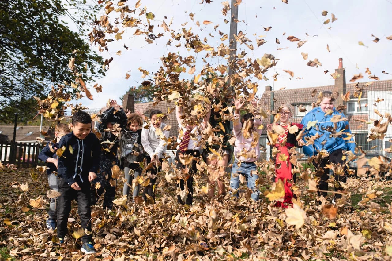 Group of Fun Fest Holiday Club children laughing as they throw a cloud of golden autumn leaves into the air on the playground - child-led outdoor play during a holiday club session