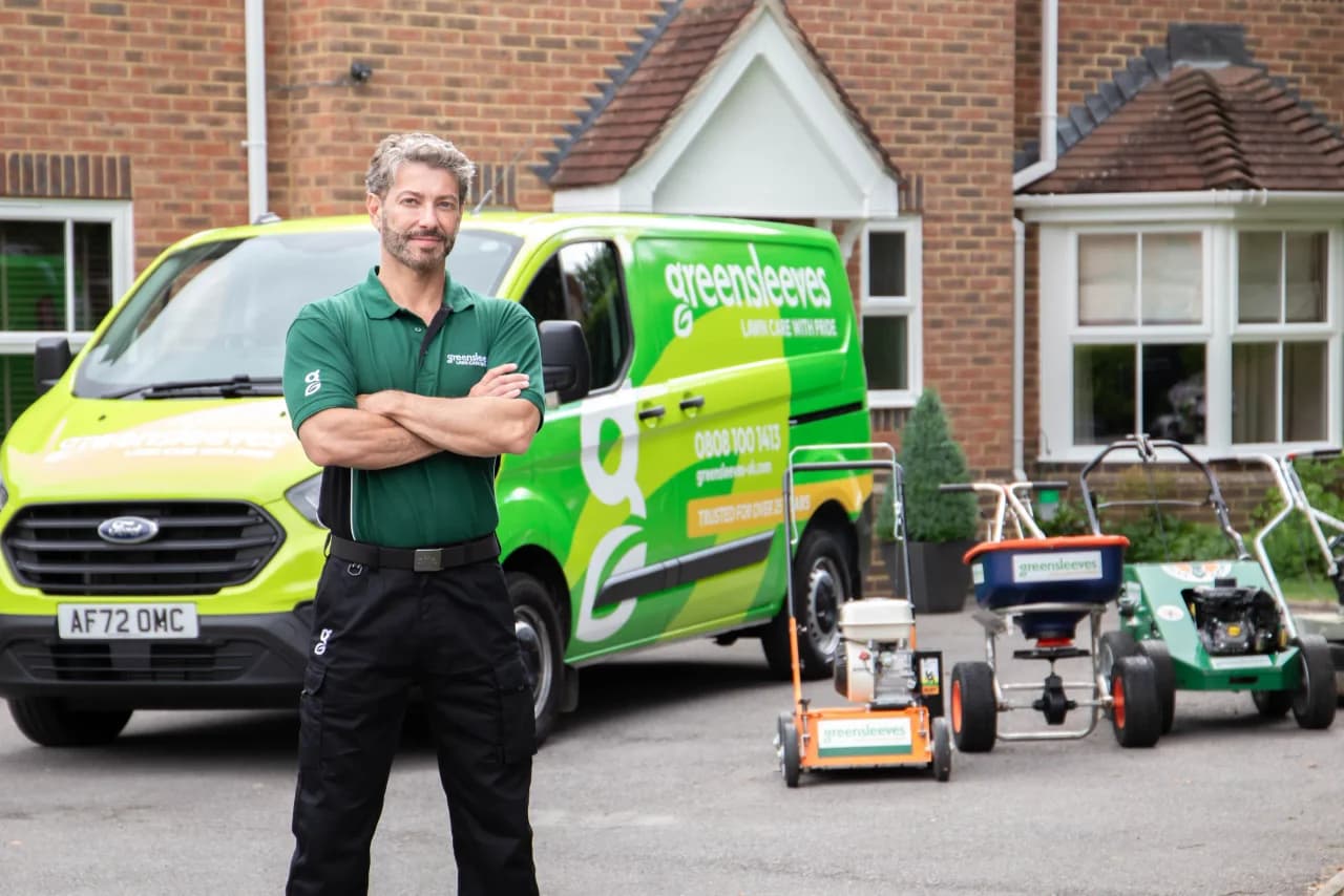 Greensleeves franchisee in branded green polo standing in front of a Greensleeves Lawn Care With Pride Ford Transit van, with three professional lawn treatment spreaders lined up alongside