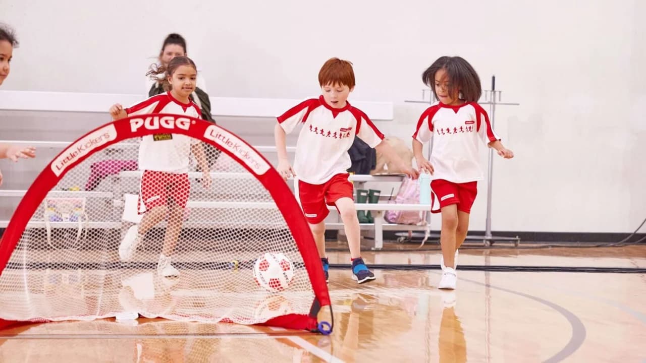 Young children in red and white Little Kickers kit playing a football game indoors with a pop-up PUGG goal in a school sports hall