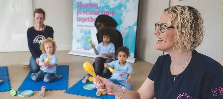 A Monkey Music teacher wearing a headset mic and holding bright-yellow maracas leads a class of mothers and toddlers sitting on blue play mats beneath a 'Sharing precious time together' brand banner