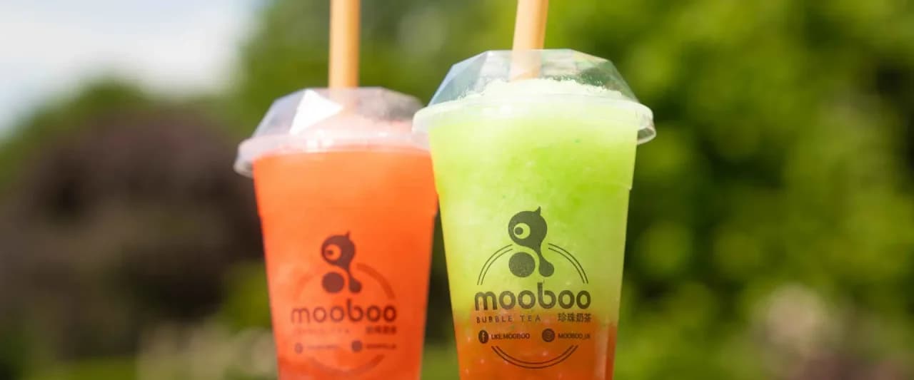 Two Mooboo Bubble Tea cups held aloft outdoors - a bright green and an orange fruit slush with bamboo straws and the Mooboo branding visible against a blurred hedge background