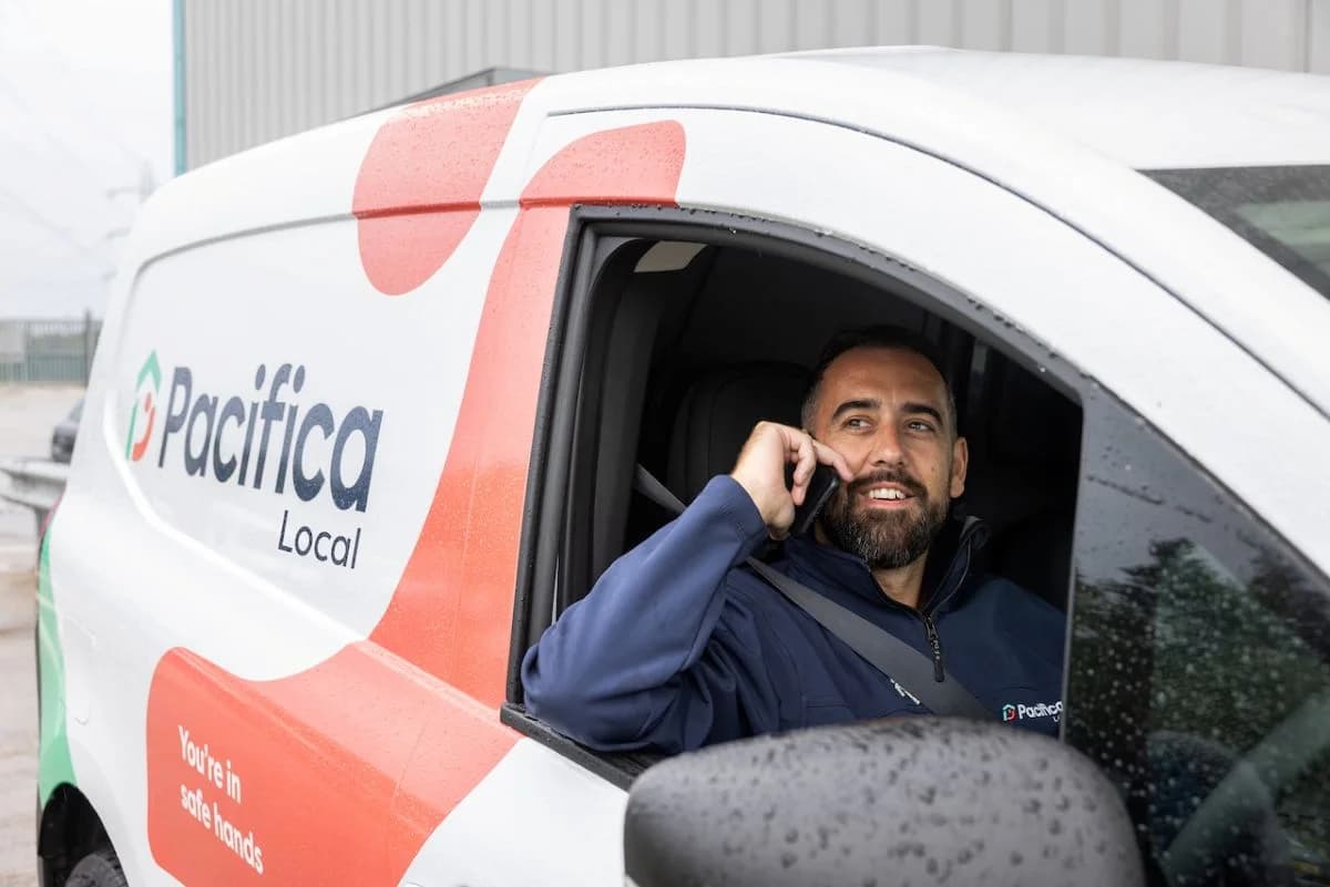 A Pacifica Local franchisee in branded navy fleece smiling on a phone call from the driver's seat of his wrapped white Pacifica Local repair van with the 'You're in safe hands' tagline visible on the side