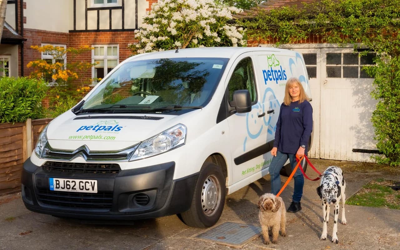 A Petpals franchisee in a branded navy fleece standing beside her white Petpals Citroën van, holding orange leads attached to a brown labradoodle and a black-and-white dalmatian on a suburban driveway
