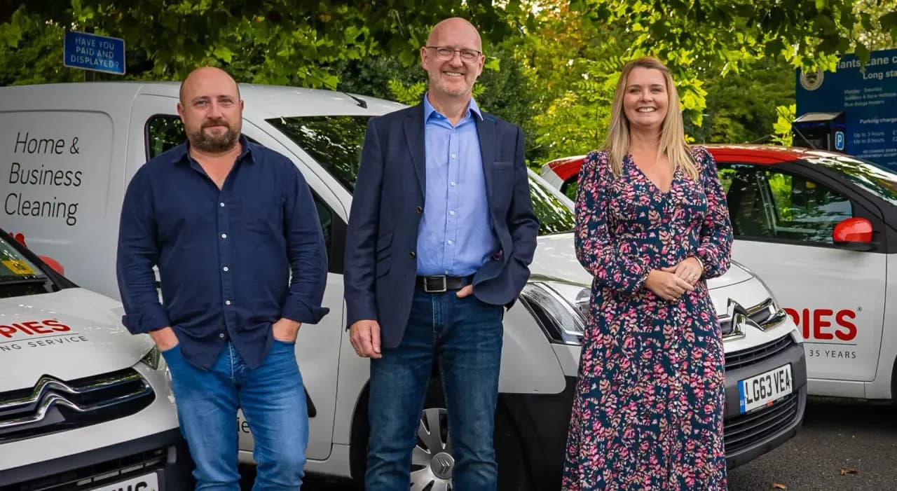 Three Poppies team members - two men in business-casual attire and a woman in a floral dress - standing in front of branded white Poppies 'Home & Business Cleaning' vans on a tree-lined street