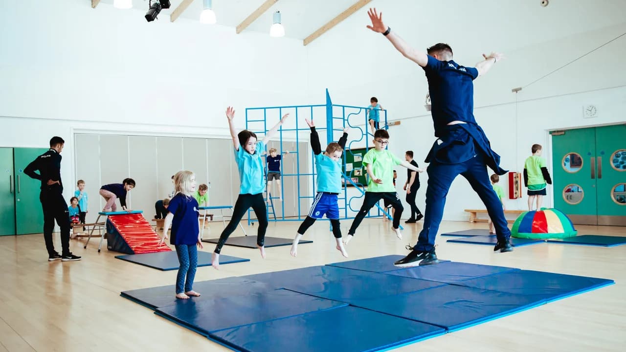 Premier Education coach in navy uniform leading a primary school PE session in a school hall, with children in blue and green PE kits jumping on blue gymnastics mats, climbing frames and a soft play tunnel in the background