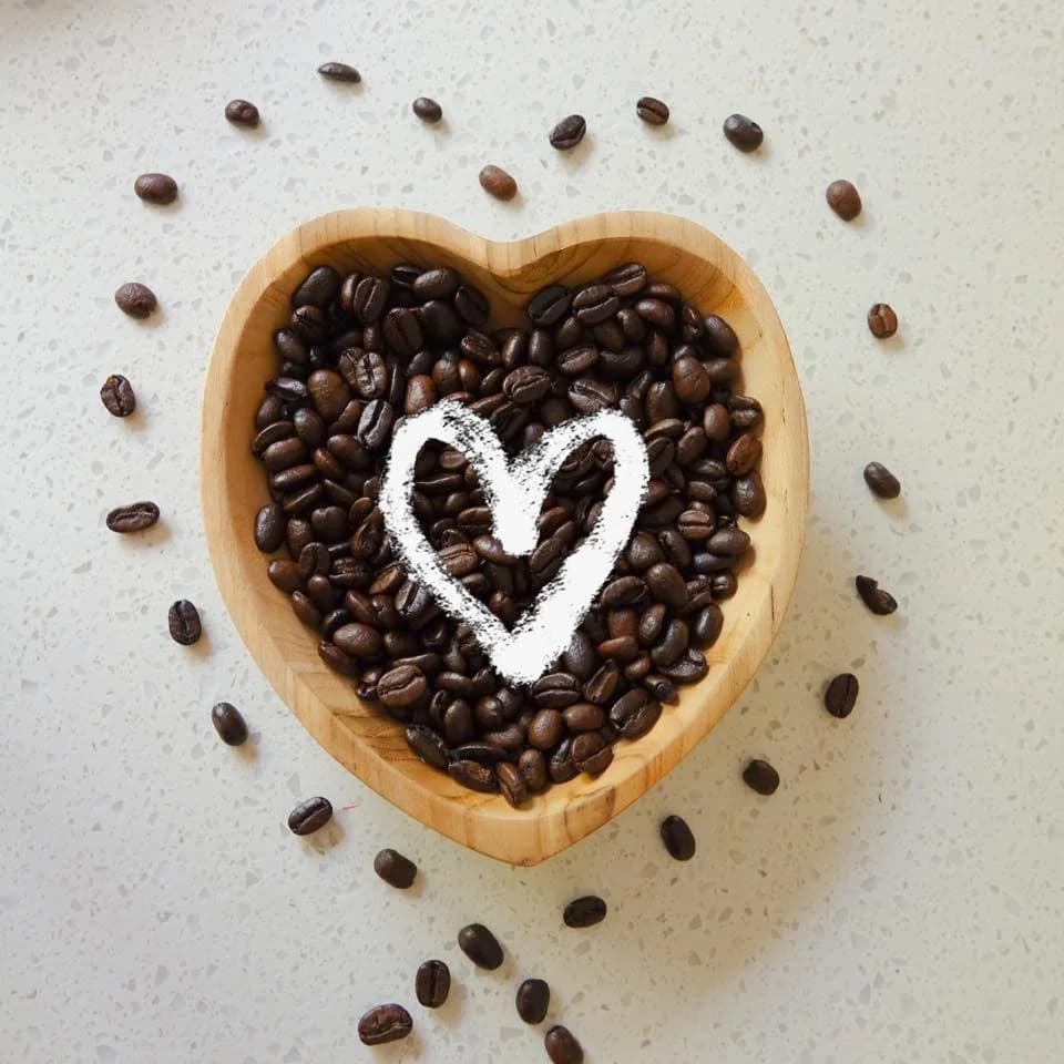 Roasted coffee beans piled inside a heart-shaped wooden bowl with a chalk heart drawn over them and loose beans scattered around on a marbled surface - Puccino's brand love-of-coffee imagery