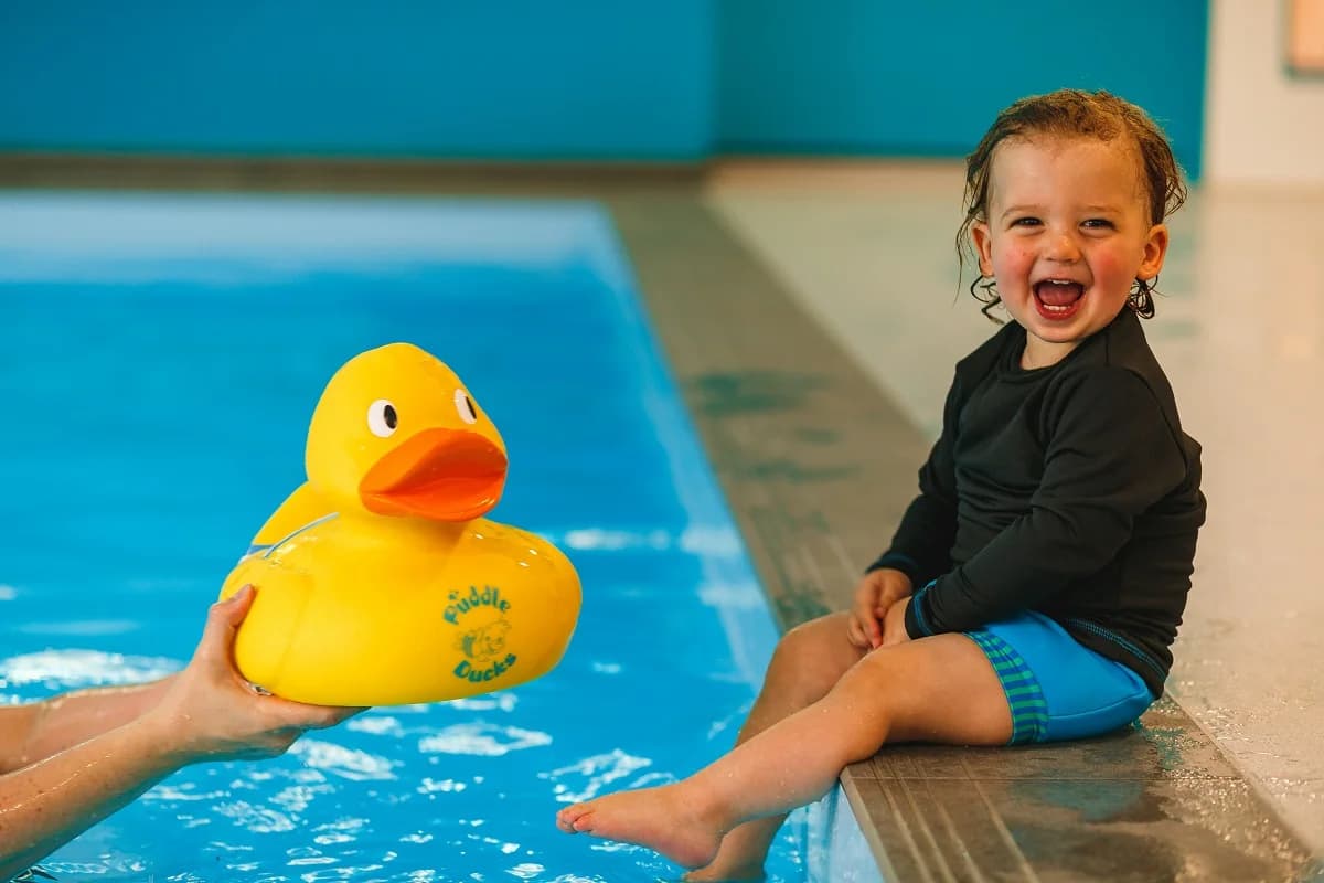 Toddler in a black and blue Puddle Ducks rash vest laughing at the edge of an indoor pool while the swim teacher holds out a yellow Puddle Ducks branded rubber duck float - the brand's child-led teaching style in action