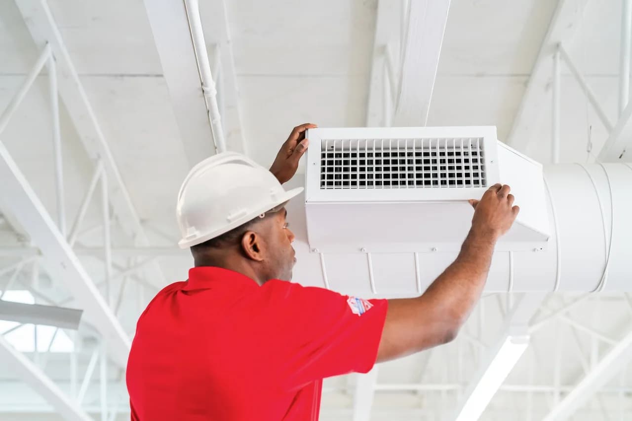 Rainbow Restoration technician in a red branded polo and white hard hat installing a white HVAC vent in a commercial ceiling, demonstrating the network's trained-engineer service delivery