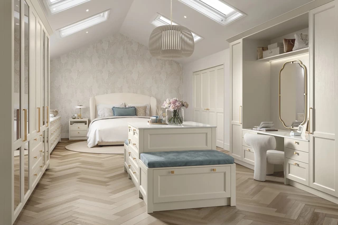 Raison Home UK cream fitted bedroom with custom wardrobes featuring gold handles, central dressing bench with blue velvet seat, dressing table corner with arched mirror, skylights overhead and herringbone wood flooring
