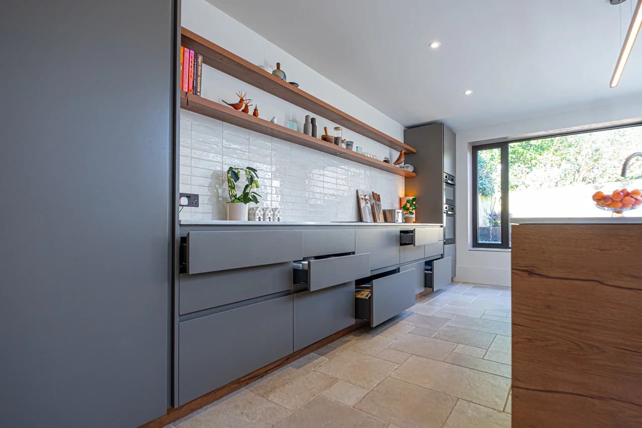 Raison Home UK modern grey handleless kitchen with open walnut shelving, white metro tile splashback, integrated appliances and a stone-tiled floor