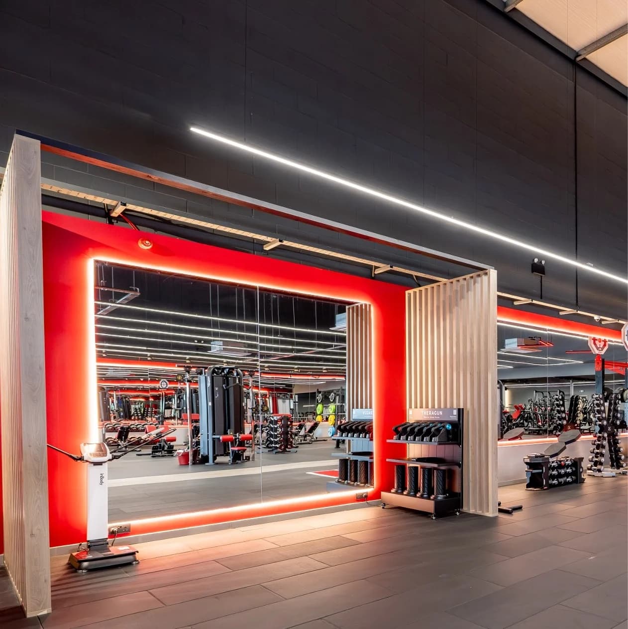 Snap Fitness club interior viewed through a wood-slatted entry - a red-accent mirror wall framed in LED strip lighting, hexagon weights rack and free-weights zone beyond, modern industrial fit-out