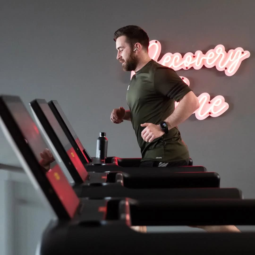 Male Snap Fitness member running on a treadmill in a green Adidas tee, with a pink neon 'Recovery' script sign glowing on the wall behind, illustrating the brand's modern cardio and recovery offering