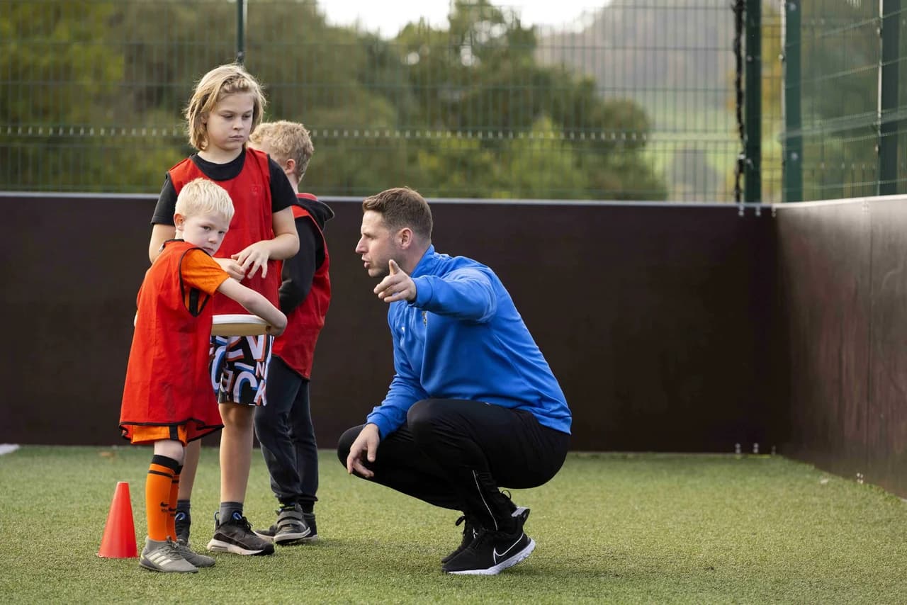 SportsCool coach in a blue branded jacket crouched on an astroturf pitch giving instructions to two children in orange and red SportsCool bibs, behind a metal mesh fence - a typical school-based SportsCool session