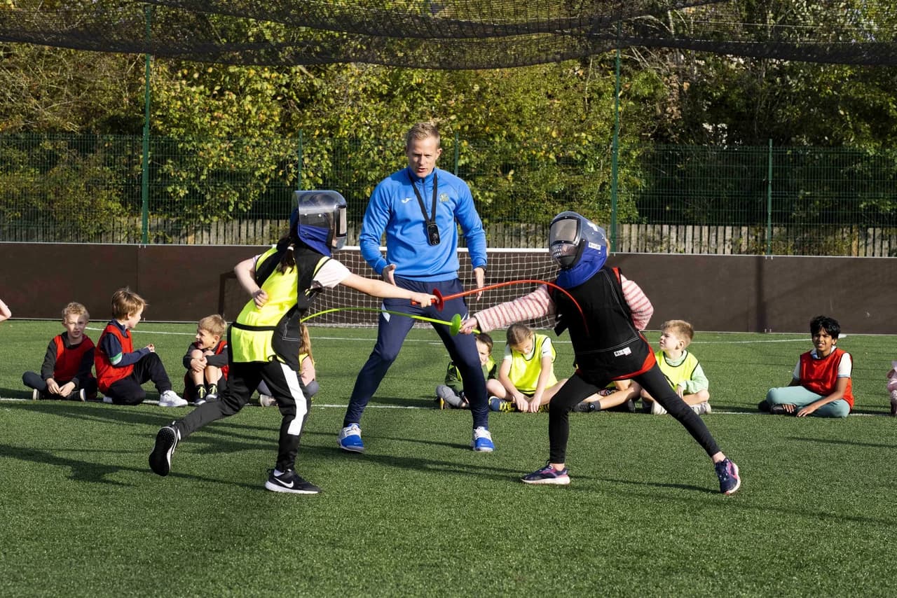Two children in fencing helmets and protective vests duelling with foam sabres on an outdoor astroturf pitch while a SportsCool coach in a blue branded top referees, with other children in red and yellow bibs watching from the sidelines