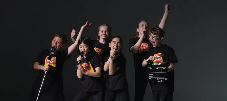 Six Stagecoach Performing Arts students in black branded tees performing animatedly with microphones and a film clapperboard against a dark grey backdrop - typical Saturday class energy