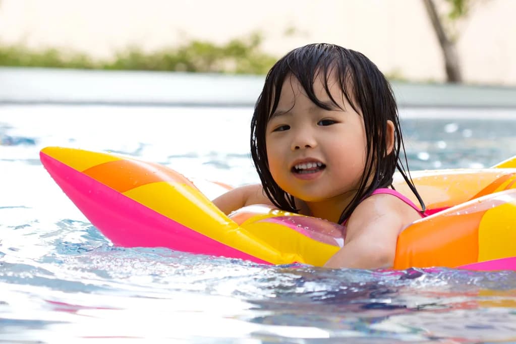 Smiling Asian toddler with wet dark hair lying on a pink, yellow and orange inflatable pool float in clear pool water - a typical confidence-in-water moment during a Swimtime lesson