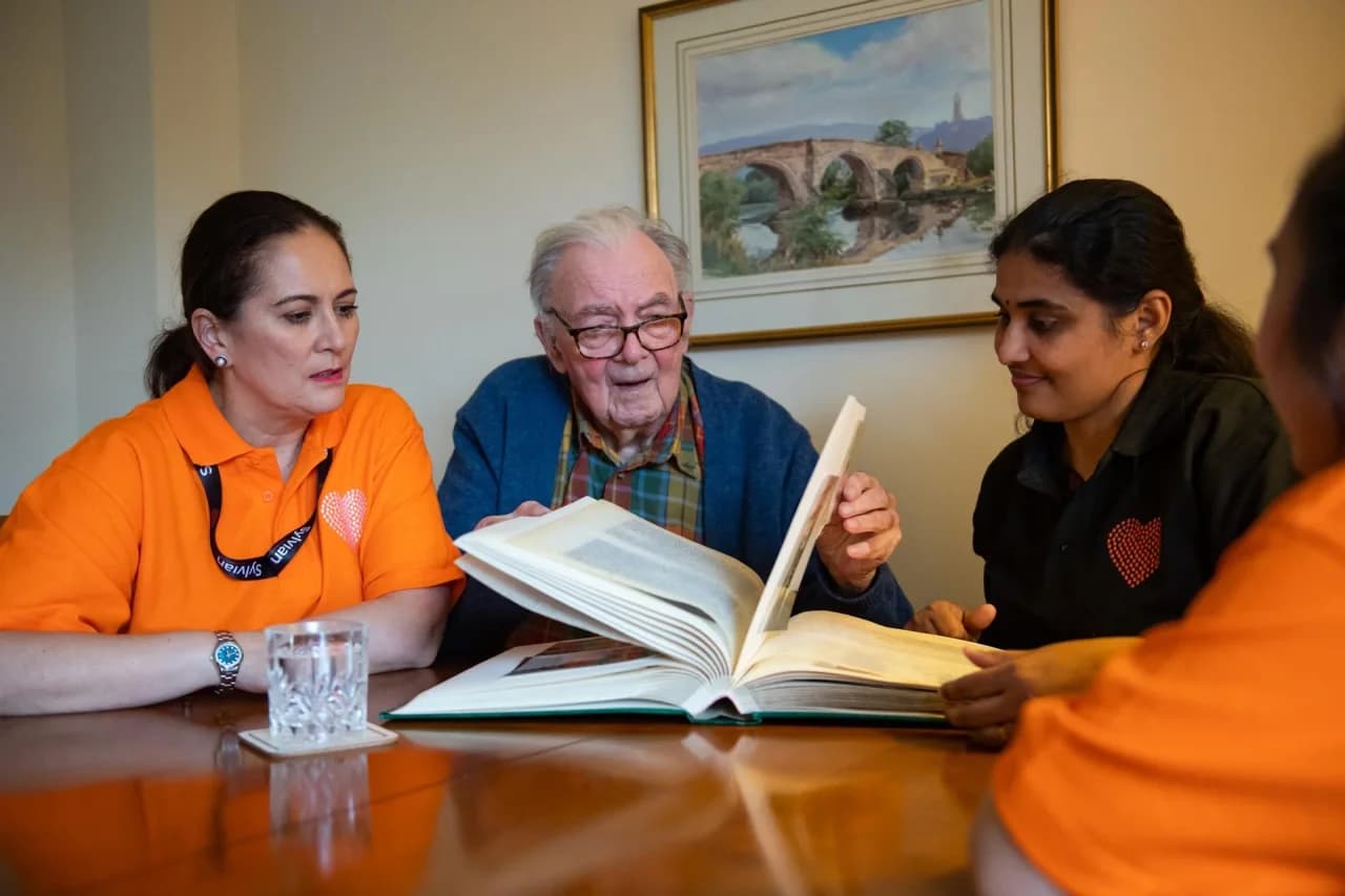 Two Sylvian Care carers - one in an orange branded polo, one in a black branded polo - sitting at a dining table with an elderly gentleman in his home looking through a hardback photo book together, a framed bridge painting on the wall behind