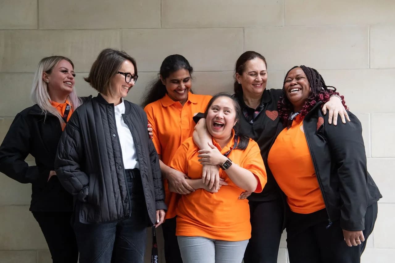 Six Sylvian Care team members in orange branded polos and black jackets laughing together against a stone wall - a typical Sylvian Care team portrait showing the people-first culture
