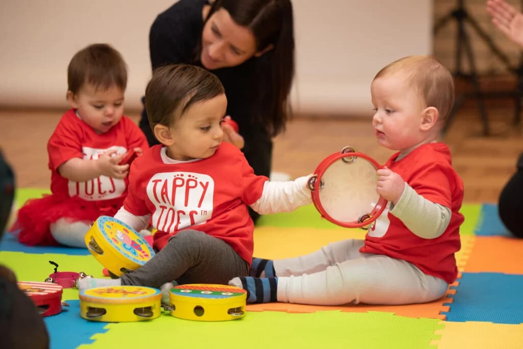 Three babies and young toddlers in red Tappy Toes tops sitting on colourful foam play mats exploring wooden tambourines, with a class instructor in the background - the youngest end of the Tappy Toes age-specific programme