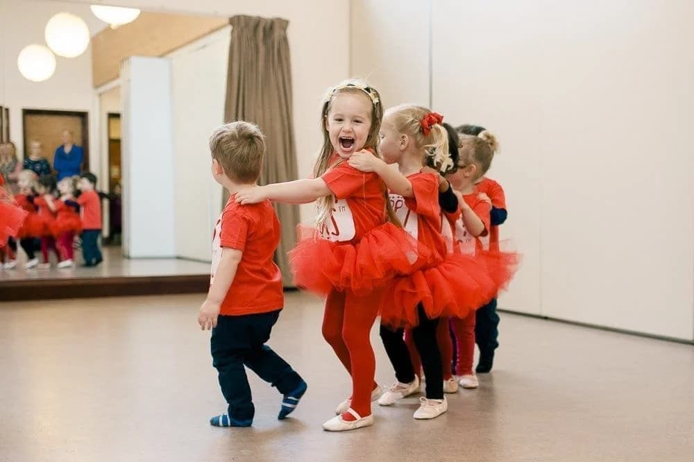 A joyful Tappy Toes class moment - preschool children in red branded tops and red tutus group-hugging mid-dance in a studio with mirrored wall, ballet shoes on a sprung dance floor