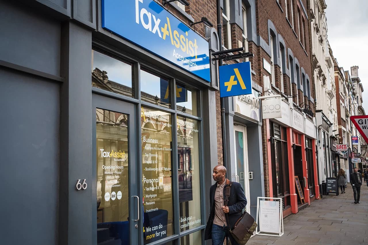 Modern TaxAssist Accountants high-street shop at 66 with full blue window signage listing business and personal services, a yellow projecting 'A+' brand sign overhead, and a man walking past on the pavement