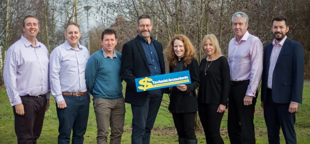 Seven TaxAssist Accountants franchisees and head-office staff standing outdoors in winter holding a TaxAssist branded sign reading 'The Accounting and Tax Service For Small Business' - illustrating the network's people and community