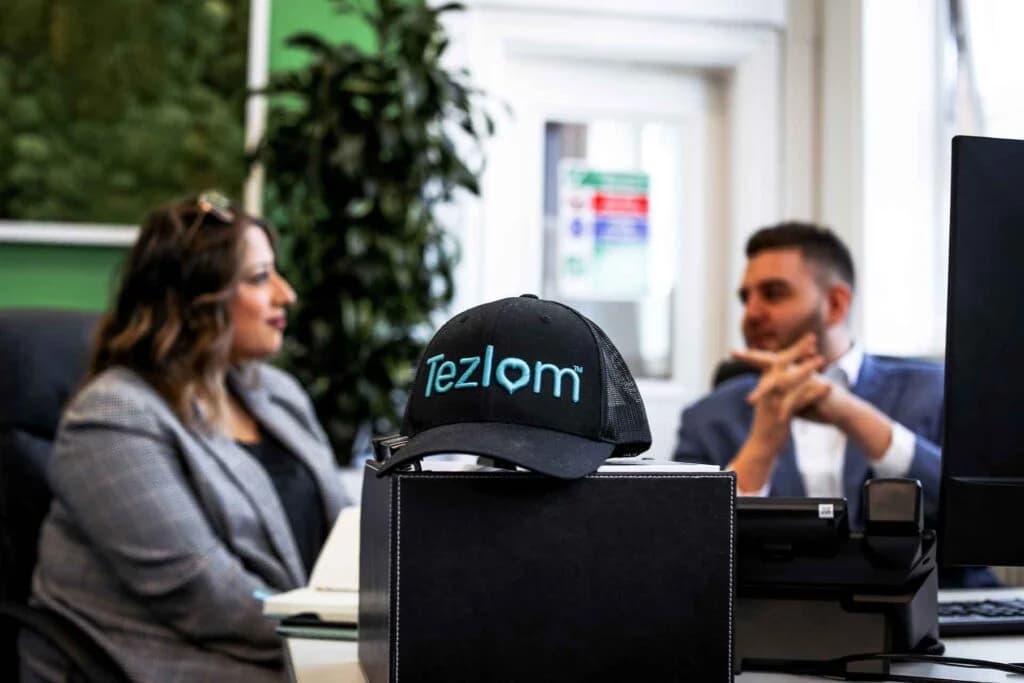 Two professionals in an office having a recruitment-style conversation across a desk, with a black Tezlom branded baseball cap in the foreground featuring the teal heart logo - illustrating the people-first culture of the network