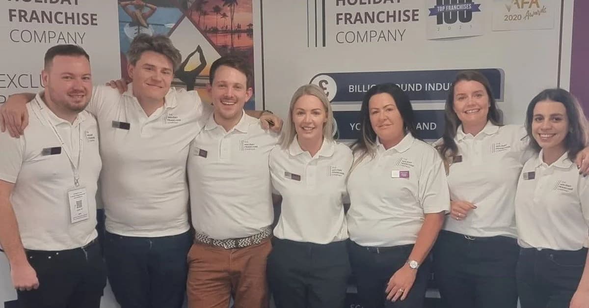 The Holiday Franchise Company team of eight in white branded polo shirts posing at a franchise exhibition stand, with the company logo and 'Top Franchises 100 2021' and 'AFA 2020 Awards' credentials displayed on the backdrop