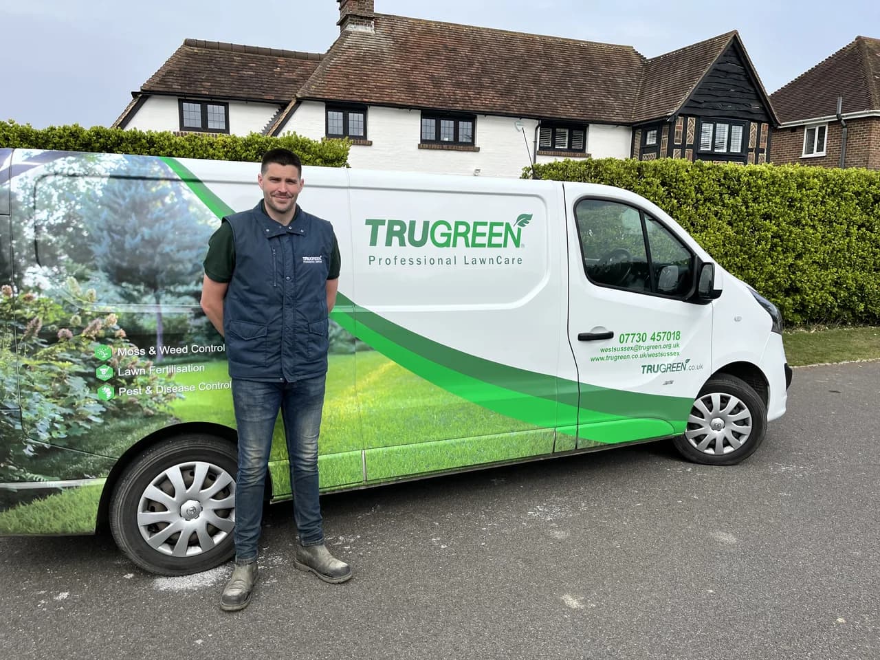A TruGreen franchisee in a branded gilet standing beside a white TruGreen Professional LawnCare van outside a tile-hung UK suburban home, the van side showing the services list (Moss & Weed Control, Lawn Fertilisation, Pest & Disease Control), a green leaf swoosh and the local 07730 457018 contact number