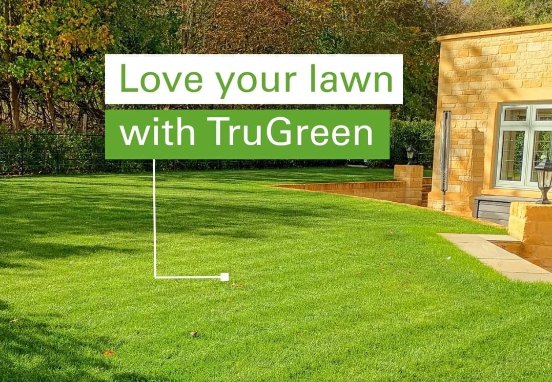 Beautifully manicured green lawn beside a Cotswold stone-built UK home, with a green-and-white marketing card reading 'Love your lawn with TruGreen' standing in the grass on a lawn-sign stake