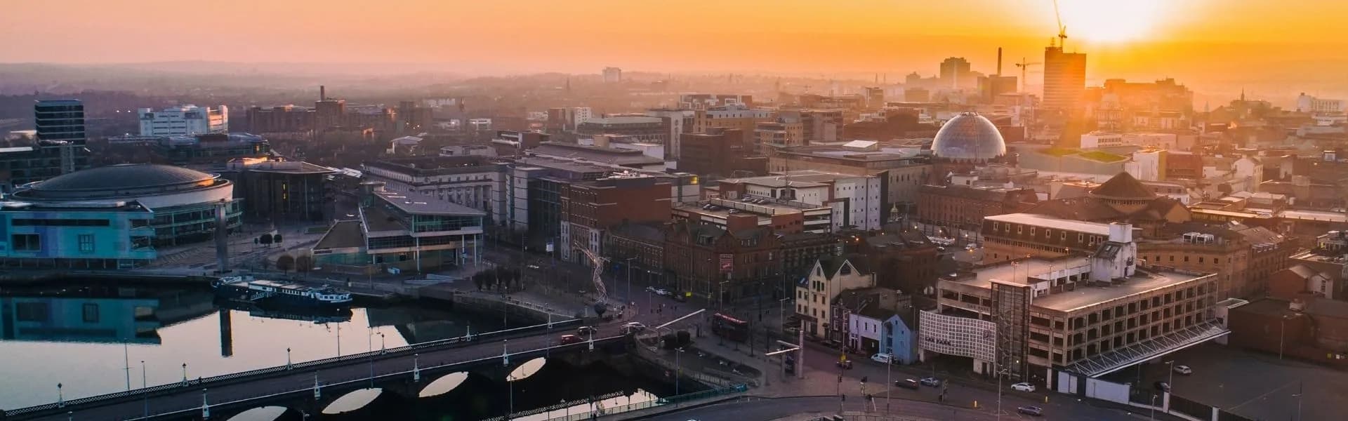 Aerial view of Belfast city centre at sunset with the River Lagan, SSE Arena and skyline silhouetted against an orange sky, representing franchise opportunities in Northern Ireland.